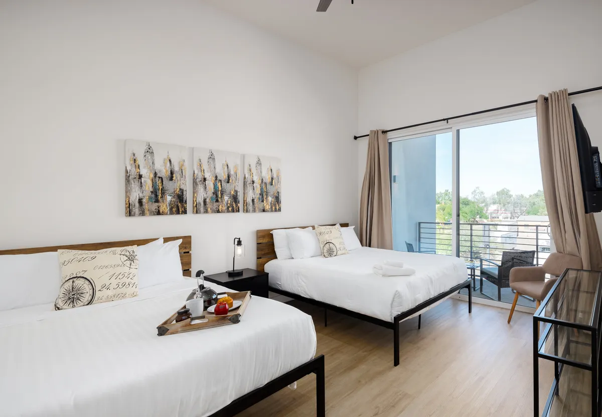 A serene bedroom scene with two beds dressed in crisp white linens, a tray with apples and a drink, and abstract wall art. The room is bathed in natural light, creating an inviting and flexible sleeping arrangement suitable for families or groups.