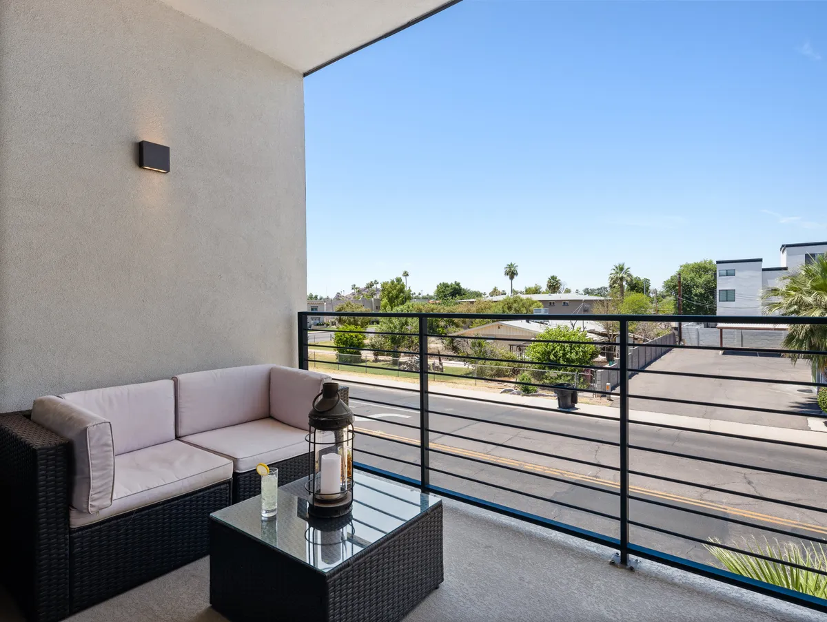 The image features an outdoor seating area with a coffee table and lantern, overlooking a street fringed with greenery. The serene atmosphere suggests an ideal space for morning coffee, informal breakout sessions, or team chats in the fresh air.