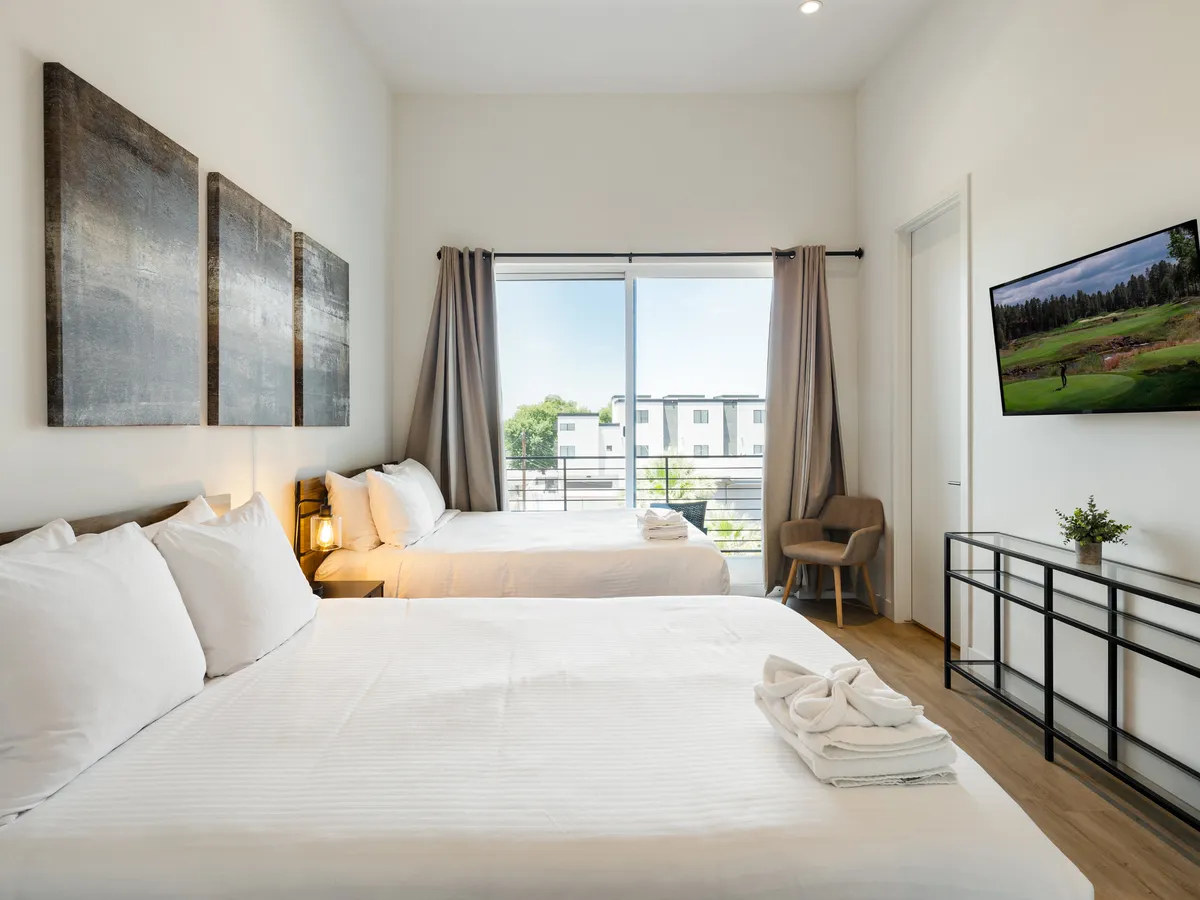 A spacious bedroom featuring two beds with white linens, a large window streaming in natural light, a wall-mounted TV, and stylish decorative art. The room radiates comfort and relaxation, hinting at the enjoyment of both restful nights and daytime lounging.
