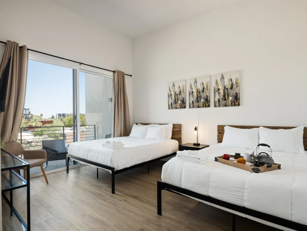 A cozy bedroom with two beds, tasteful wall art, and a view of the balcony outside. The setting is bright and inviting, allowing for shared moments or peaceful alone time, ideal for families or groups.