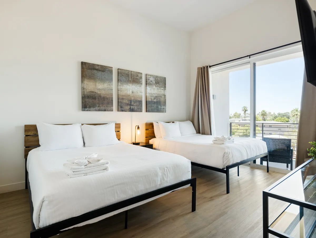 A serene bedroom with two beds, elegant artwork on the wall, and a balcony view visible through sliding doors. The room is tranquil, perfect for relaxation, and offers guests a chance to step out onto a private balcony for fresh air or a morning coffee.