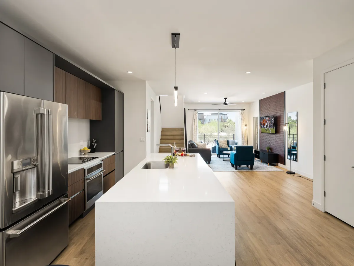 A contemporary open-plan kitchen and living space featuring a stylish kitchen island, stainless steel appliances, blue accent chairs in the living area, large windows letting in natural light, and a staircase leading to an upper level. The space is uncluttered and functional, designed for both relaxation and entertaining.