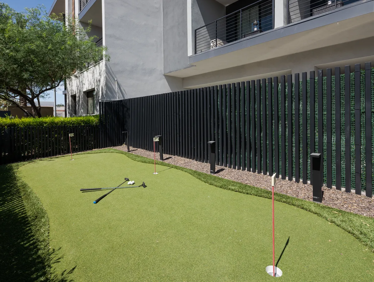 A bright outdoor putting green bordered by tasteful landscaping and a decorative fence. Golf clubs and seating areas invite residents to practice their swing, socialize, or simply enjoy the sunny Arizona climate.