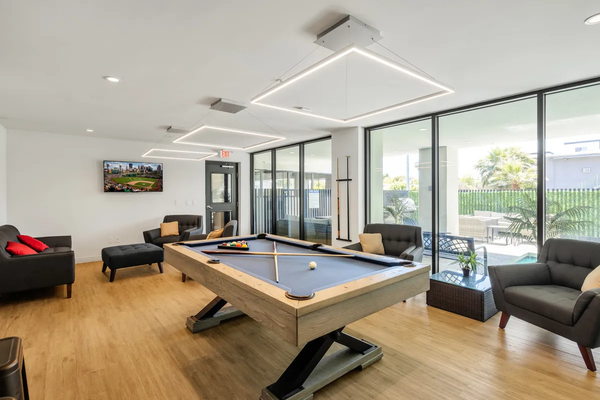 A spacious, modern clubhouse interior featuring a pool table, comfortable sofas, large television, and floor-to-ceiling windows. The setting is ideal for group relaxation, games, and unwinding after a night out.